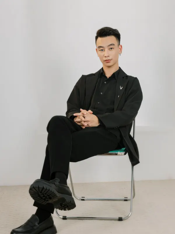 Jeune homme mince en surchemise et pantalon noirs sur une chaise en métal, studio épuré