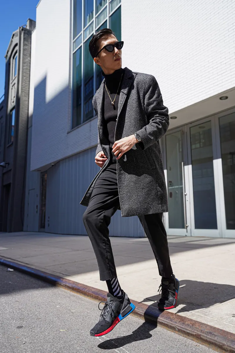Jeune homme mince en manteau de laine gris et pantalon noir slim en pleine marche dans une rue ensoleillée