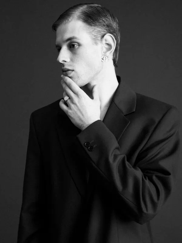Portrait noir et blanc d’un jeune homme élancé en veste de costume sombre, main au menton