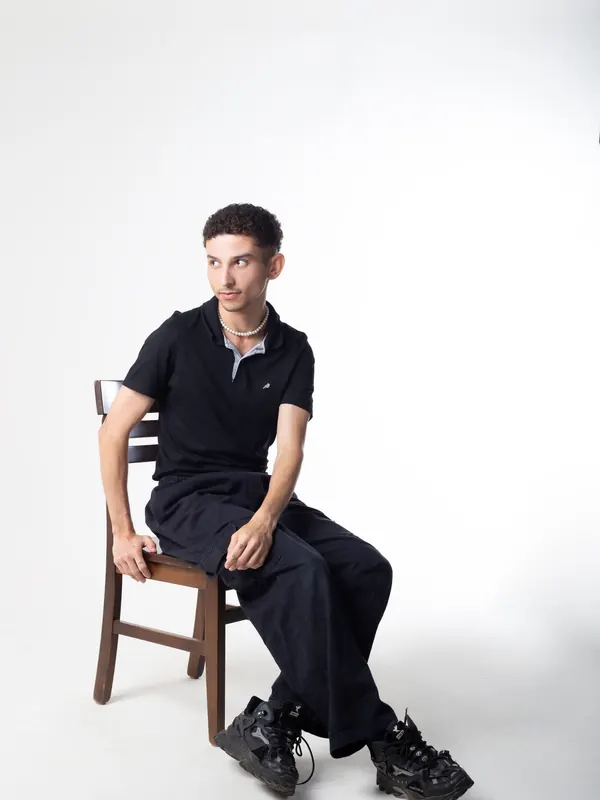 Jeune homme très mince en polo marine et pantalon sombre assis sur une chaise, studio lumineux