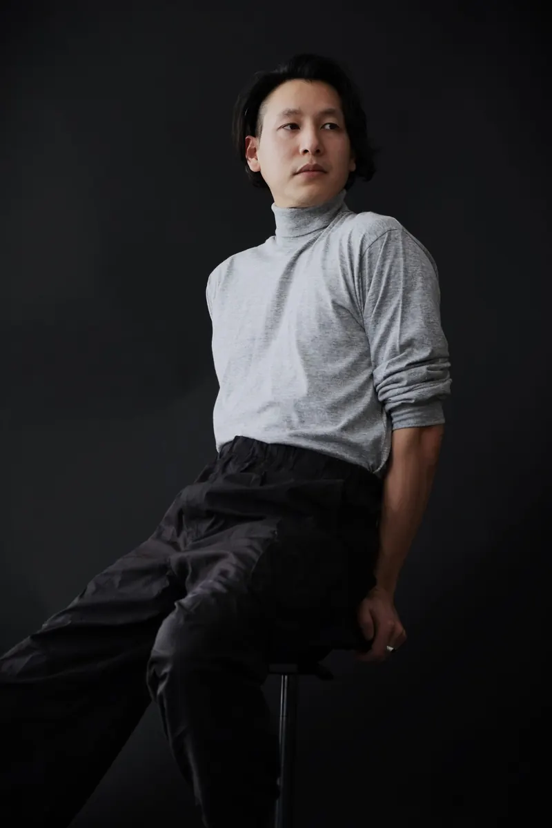 Slender young man in a grey roll-neck and black trousers against a dark studio wall