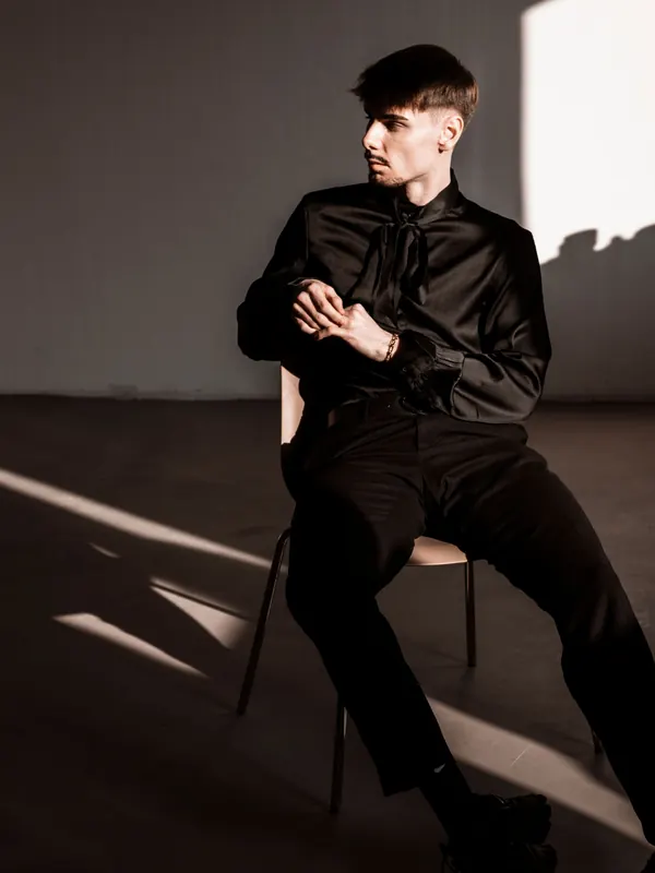 Jeune homme mince en chemise et pantalon noirs assis dans une lumière de fenêtre dure et des ombres