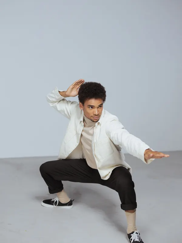 Jeune homme mince en veste crème et pantalon noir court dans une pose dynamique en studio
