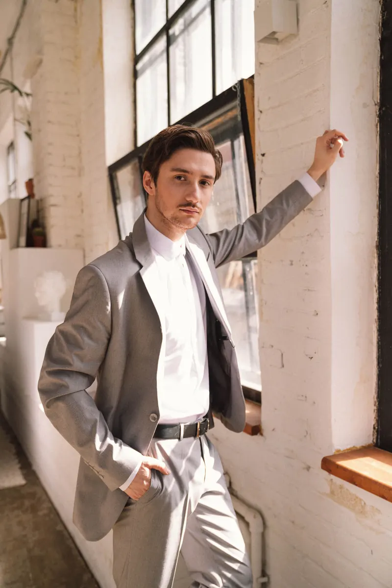 Slender young man in a grey suit and white shirt posing by an industrial window